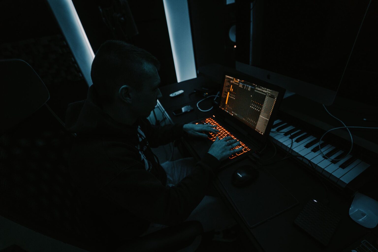 Mężczyzna w ciemnym studio muzycznym pracuje na laptopie z podświetlaną klawiaturą, tworząc muzykę hardstyle