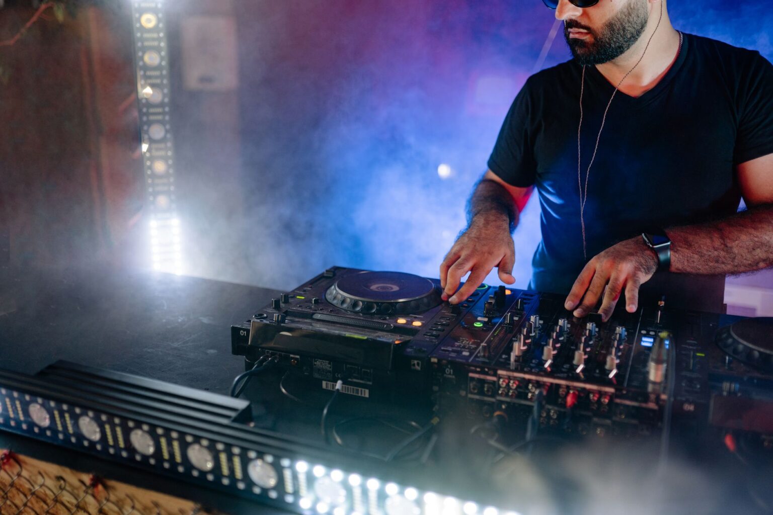 Brodaty mężczyzna w czarnej koszulce obsługuje profesjonalny sprzęt DJ w klubowym świetle i dymie
